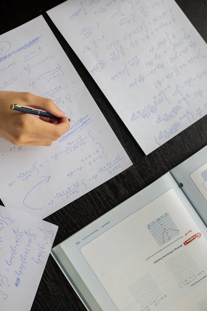 Close-up of a student solving mathematical equations with papers and textbook on a table.